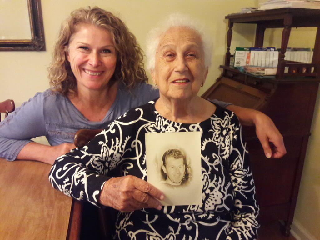 daughter sitting with her gray-haired, widowed mother holding a photo of her deceased husband as a young man in the military